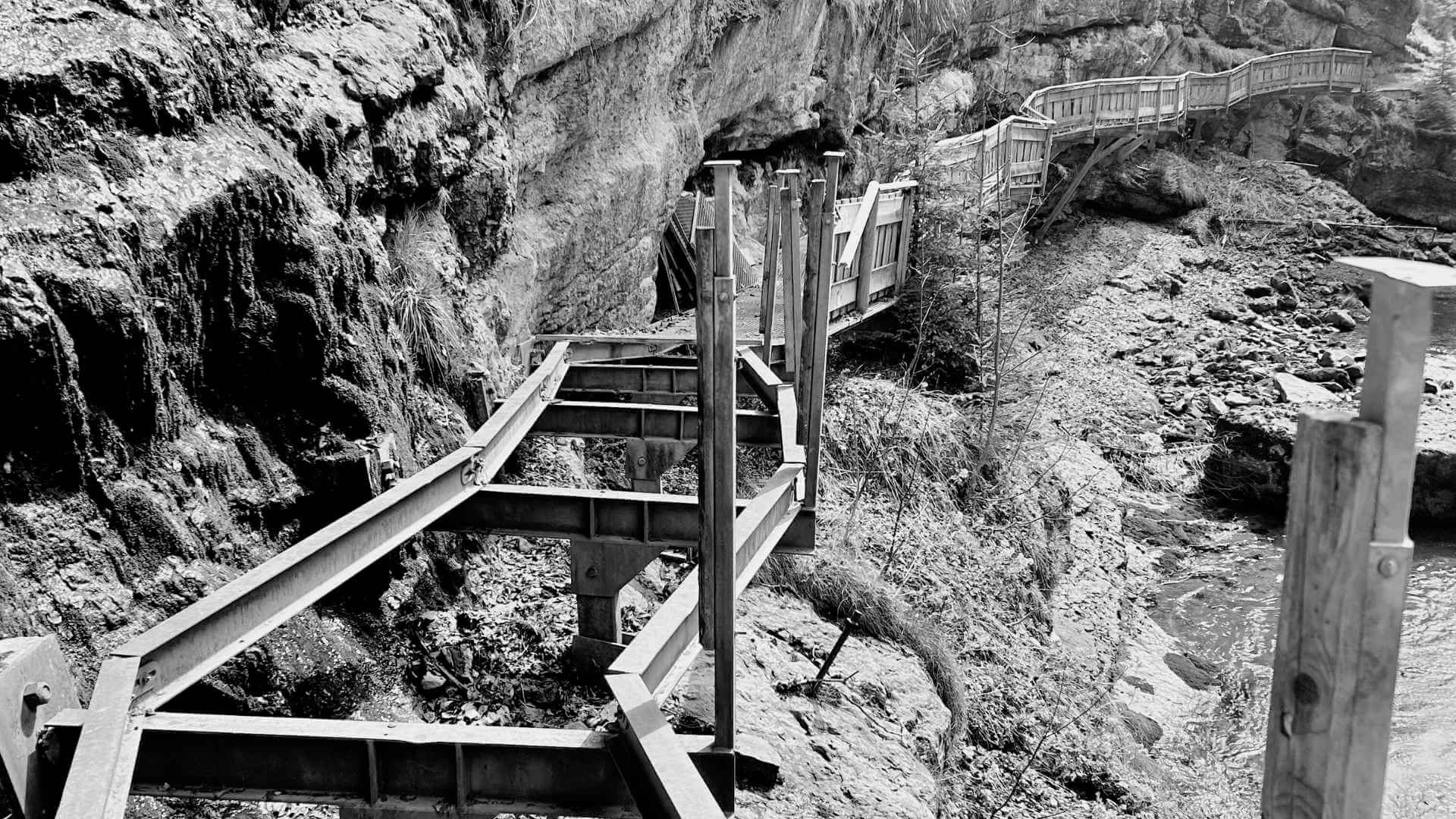 Schwarz-weiß-Foto eines zerstörten Brückenteils in einer Schlucht. Die Stahlträger sind noch da, aber die Planken fehlen – der Weg ist unterbrochen.